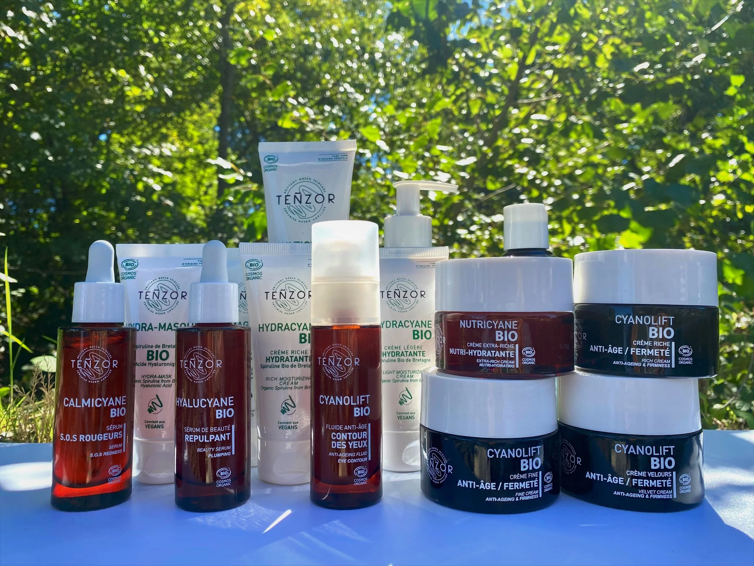 Collection de produits cosmétiques biologiques Tenzor avec crèmes, sérums et masques, disposés en plein air sous les arbres.
