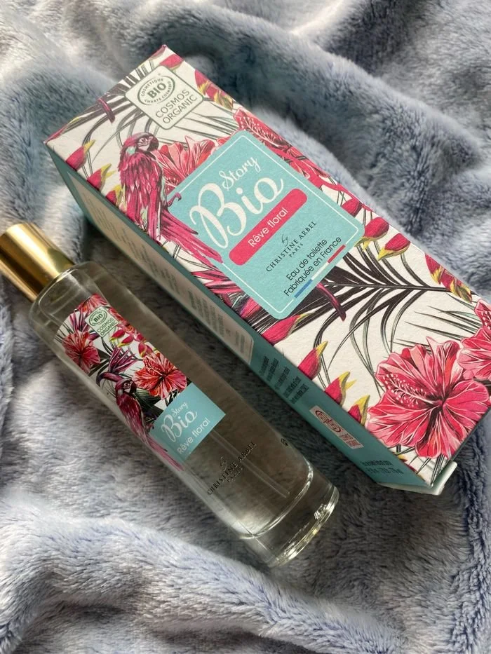 Une boîte et un flacon de parfum Bio Rêve floral de la marque Christophe Robin, avec des motifs de fleurs et de feuillage rouges et roses sur l'emballage, reposant sur une couverture en tissu gris.