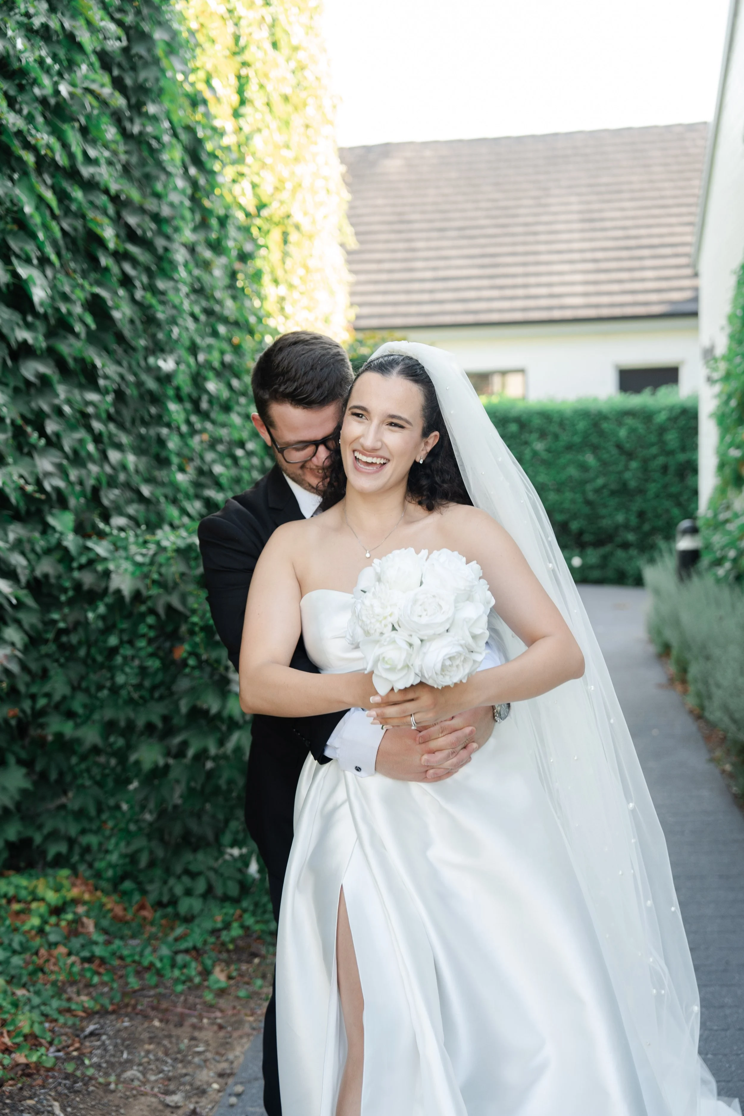 Bride and groom on their wedding day, embracing outdoors on a pathway, surrounded by greenery.