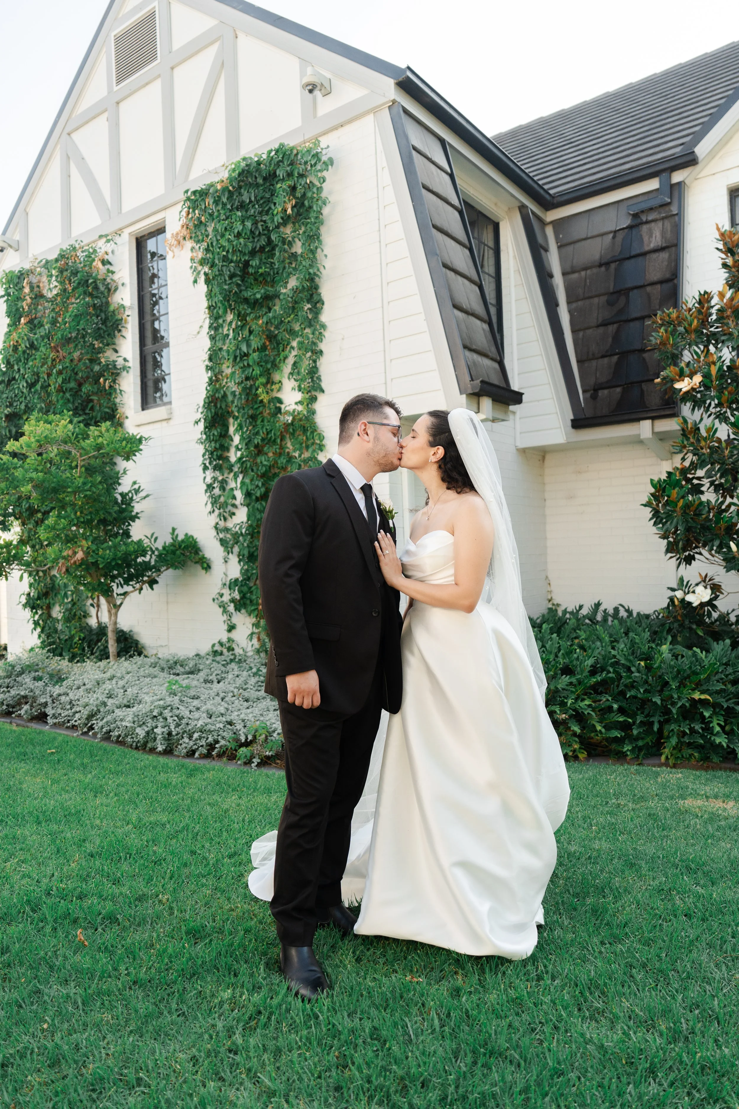 A bride and groom kissing outside in front of a white house with greenery.
