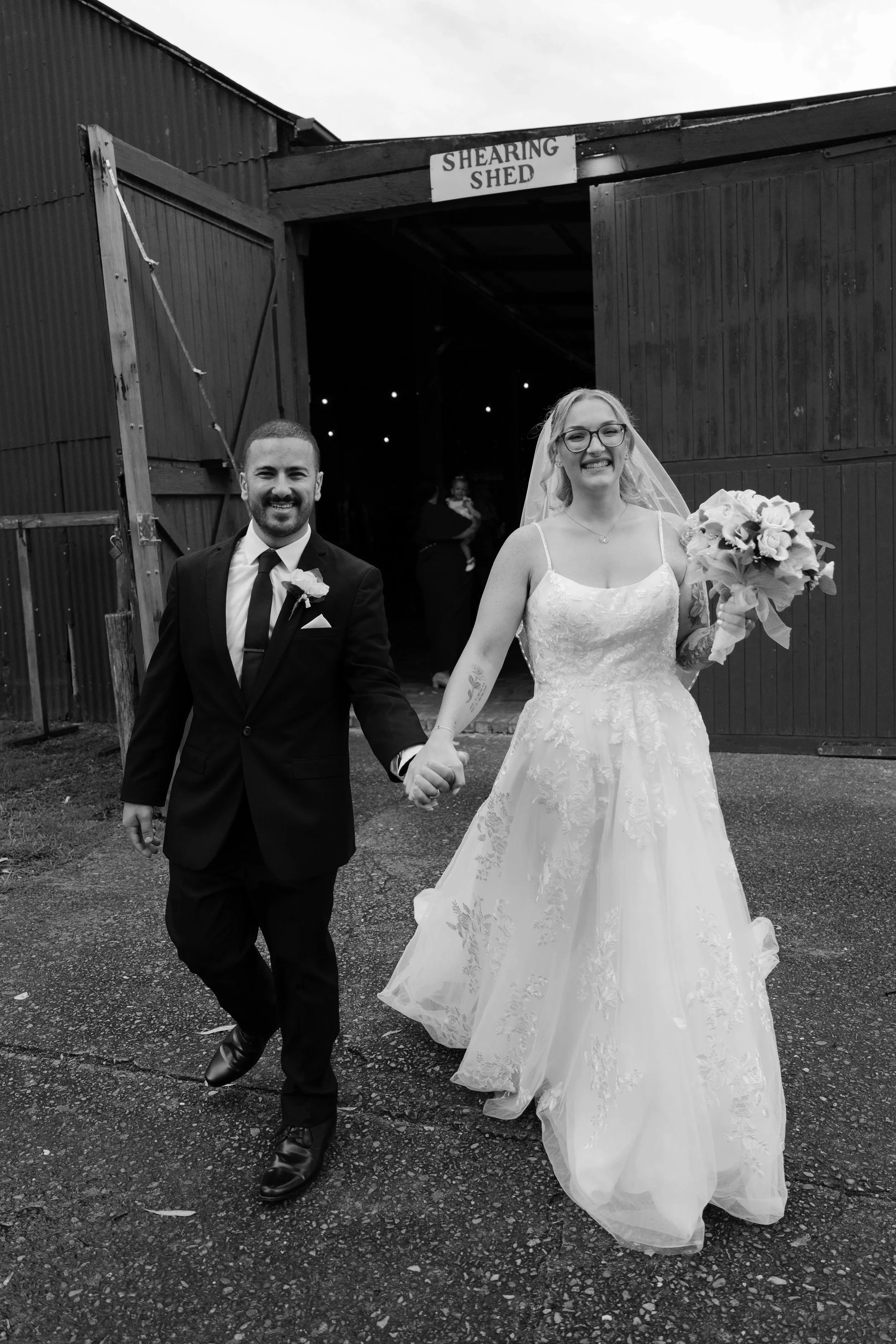 A newly married couple holding hands and walking out of a barn labeled 'Shearing Shed'. The bride is in a white wedding dress holding a bouquet, and the groom is in a black suit with a tie. At Gledswood Homestead & Winery. 