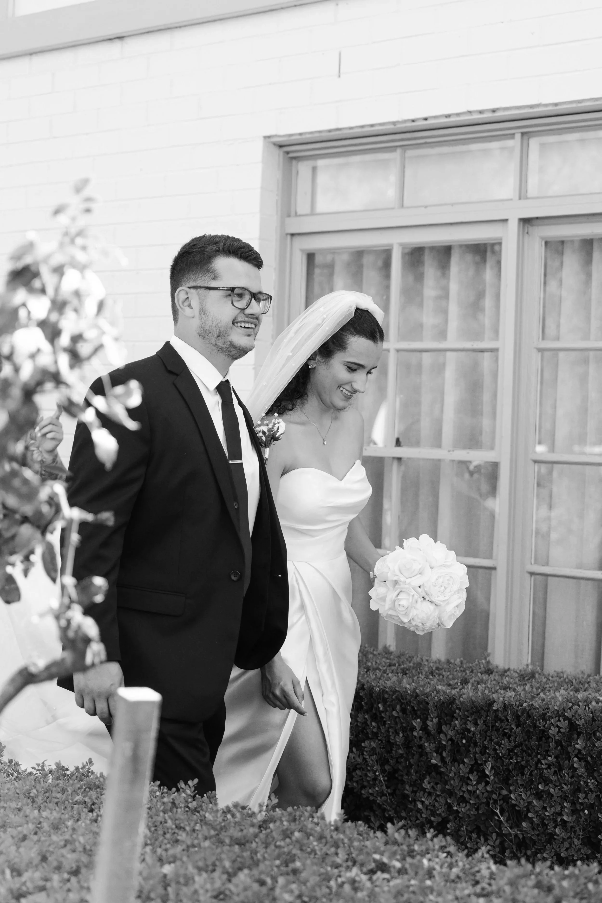 Black and white photo of a newlywed couple walking outside. The groom is in a suit with glasses, and the bride is in a strapless wedding dress with a veil, holding a bouquet of roses, smiling. At Camden Valley Inn. 