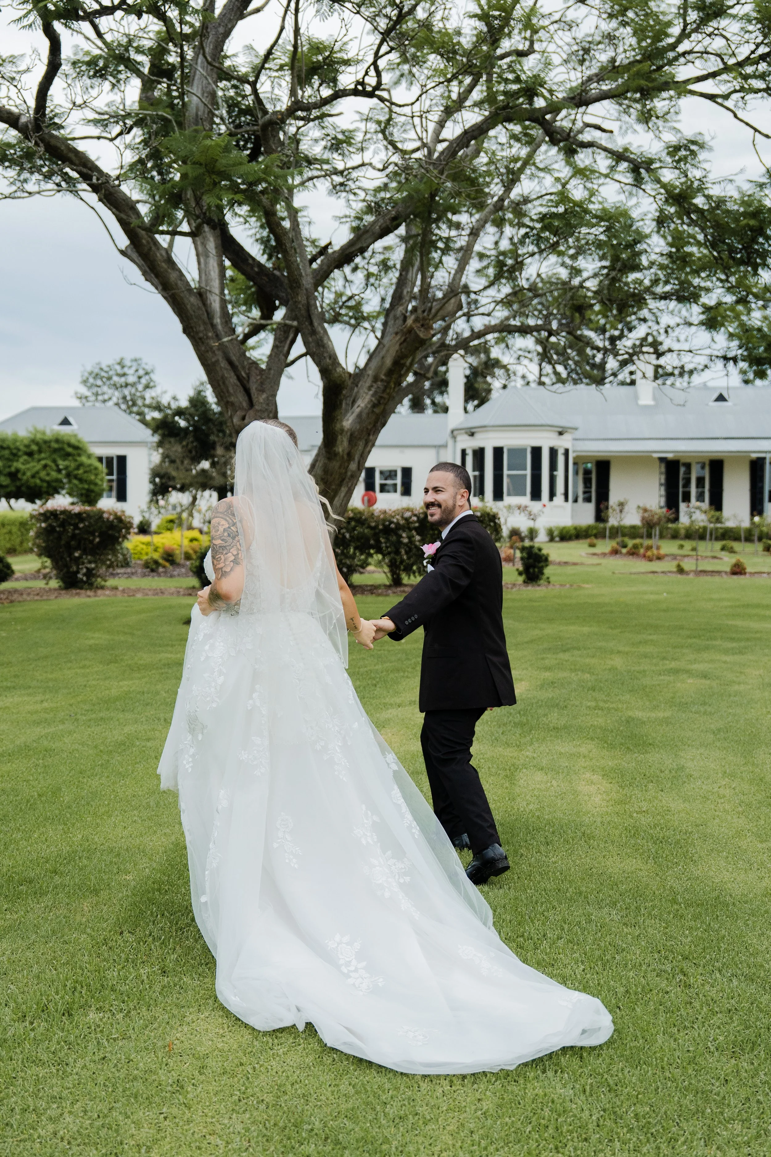 A bride and groom holding hands in a grassy yard with a large tree and white house in the background, during a wedding. At Gledswood Homestead. & Winery.