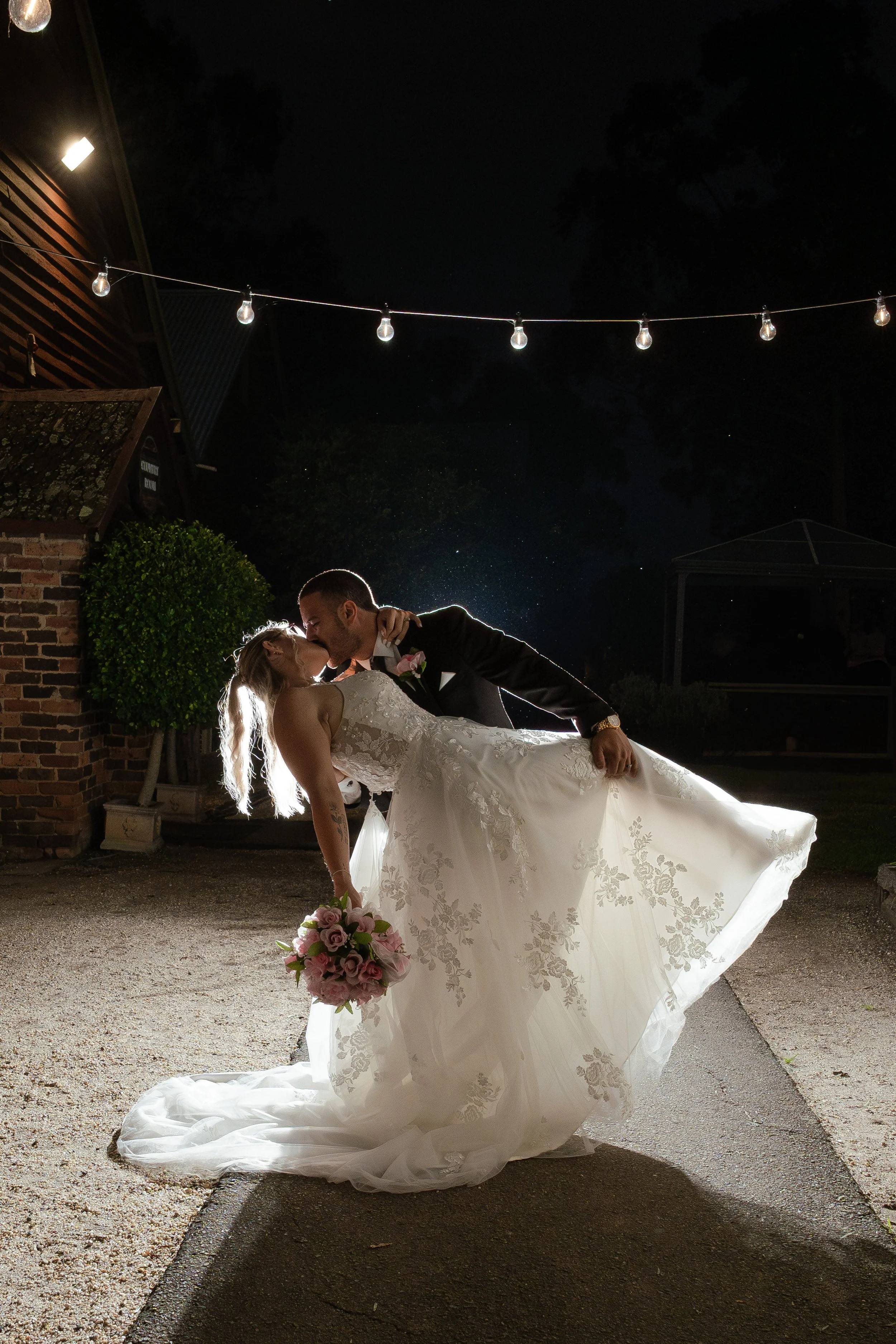 A newlywed couple sharing a kiss outdoors at night. The groom dips the bride who is holding a bouquet of pink and white flowers, with string lights overhead illuminating the scene. At Gledswood Homestead & Winery. 