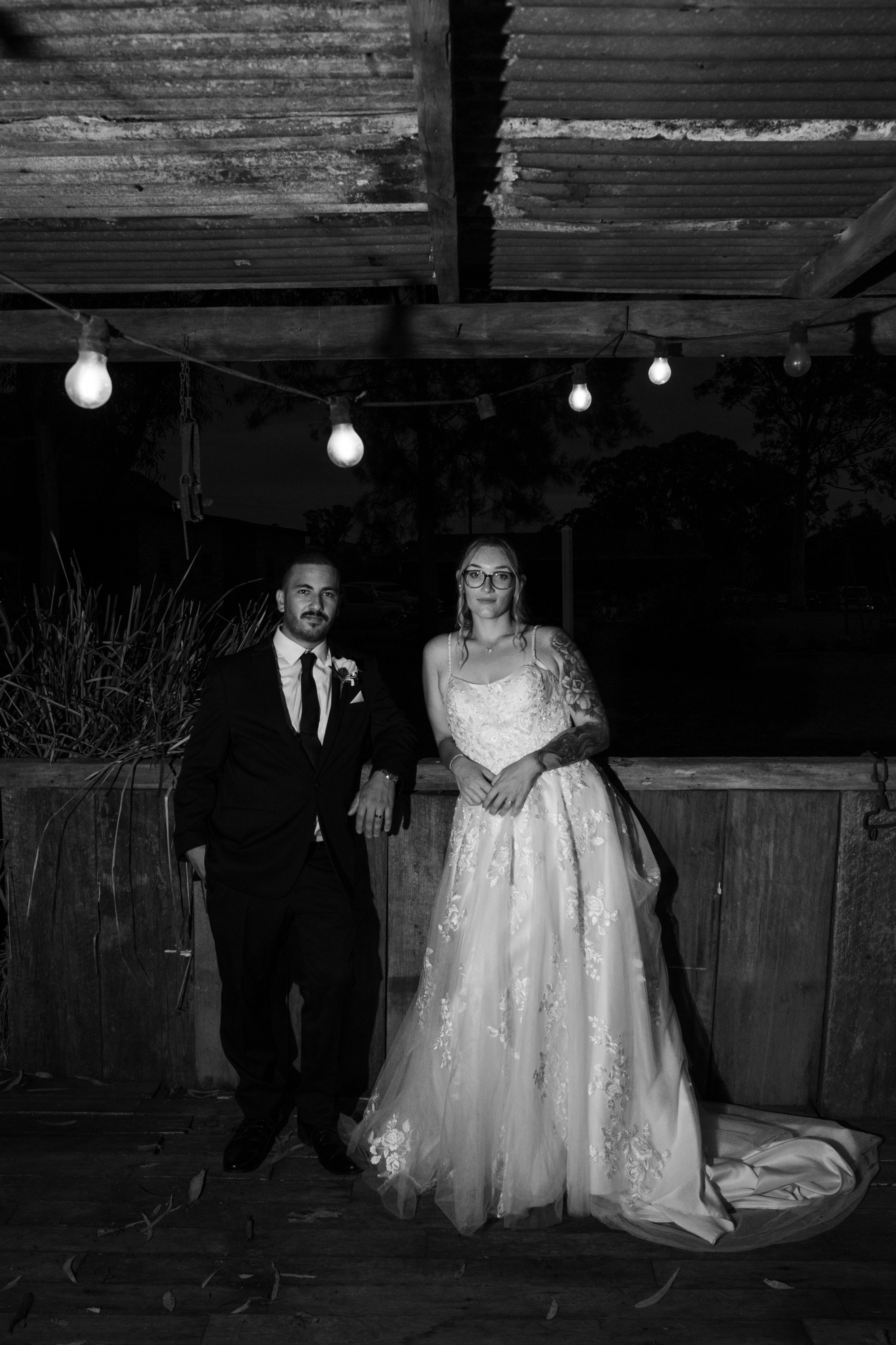 A couple in wedding attire posing outdoors at night with string lights overhead, the man in a suit and the woman in a wedding dress, standing against a wooden fence. At Gledswood Homestead & Winery.