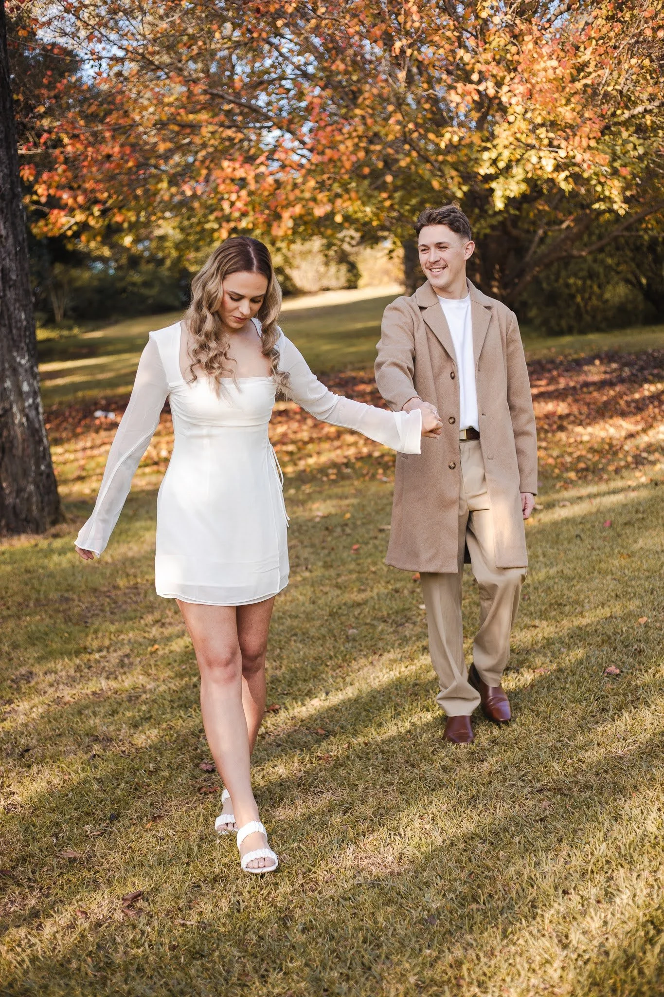 A man and woman walking outdoors on a grassy field with colorful autumn trees in the background. The woman is wearing a white dress and white sandals, and the man is wearing a beige coat and brown shoes. They are holding hands, and the woman appears to be looking down while the man is smiling.
