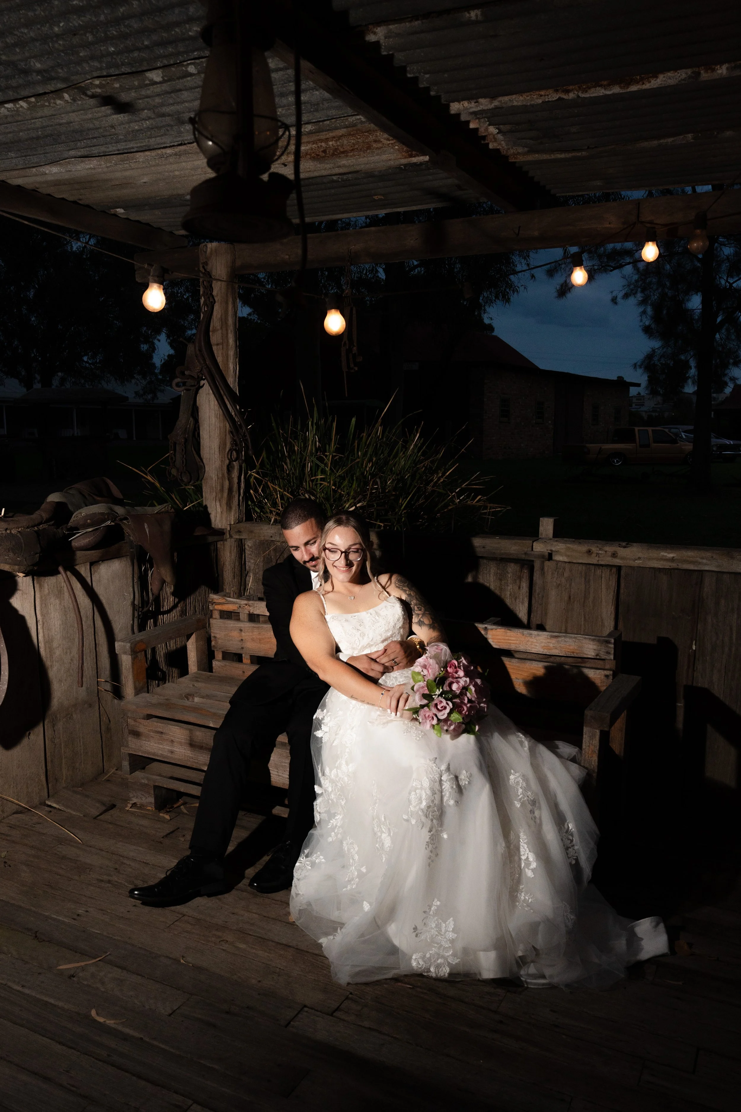 A newlywed couple sitting on a wooden bench at night. The bride is wearing a white wedding dress and holding a bouquet of pink and purple flowers; the groom is in a black suit. They are smiling and embracing. At Gledswood Homestead & Winery.