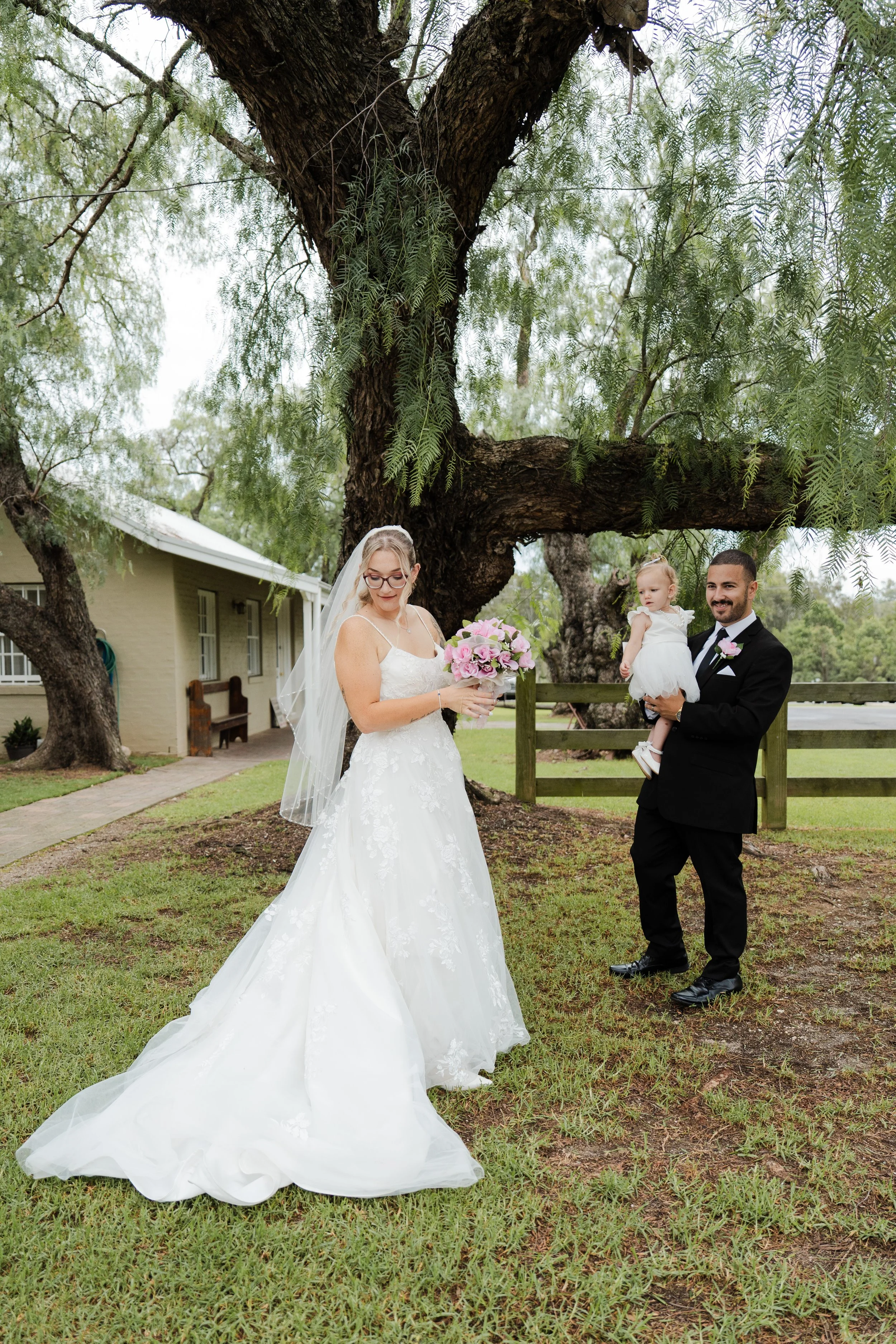 Bride in a white wedding dress holding a bouquet of pink flowers, standing under a large tree. Groom in a black tuxedo holding a young girl dressed in white, with a house and green landscape in the background. At Gledswood Homestead & Winery. 