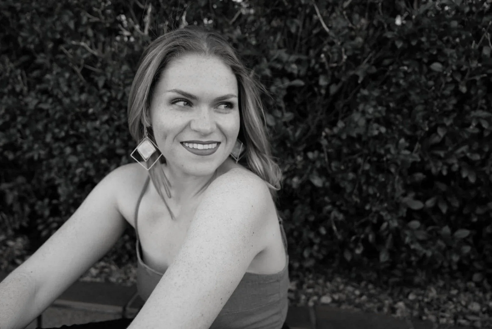 A woman with shoulder-length hair and large geometric earrings, smiling and looking to the side, outdoors with bushes in the background.