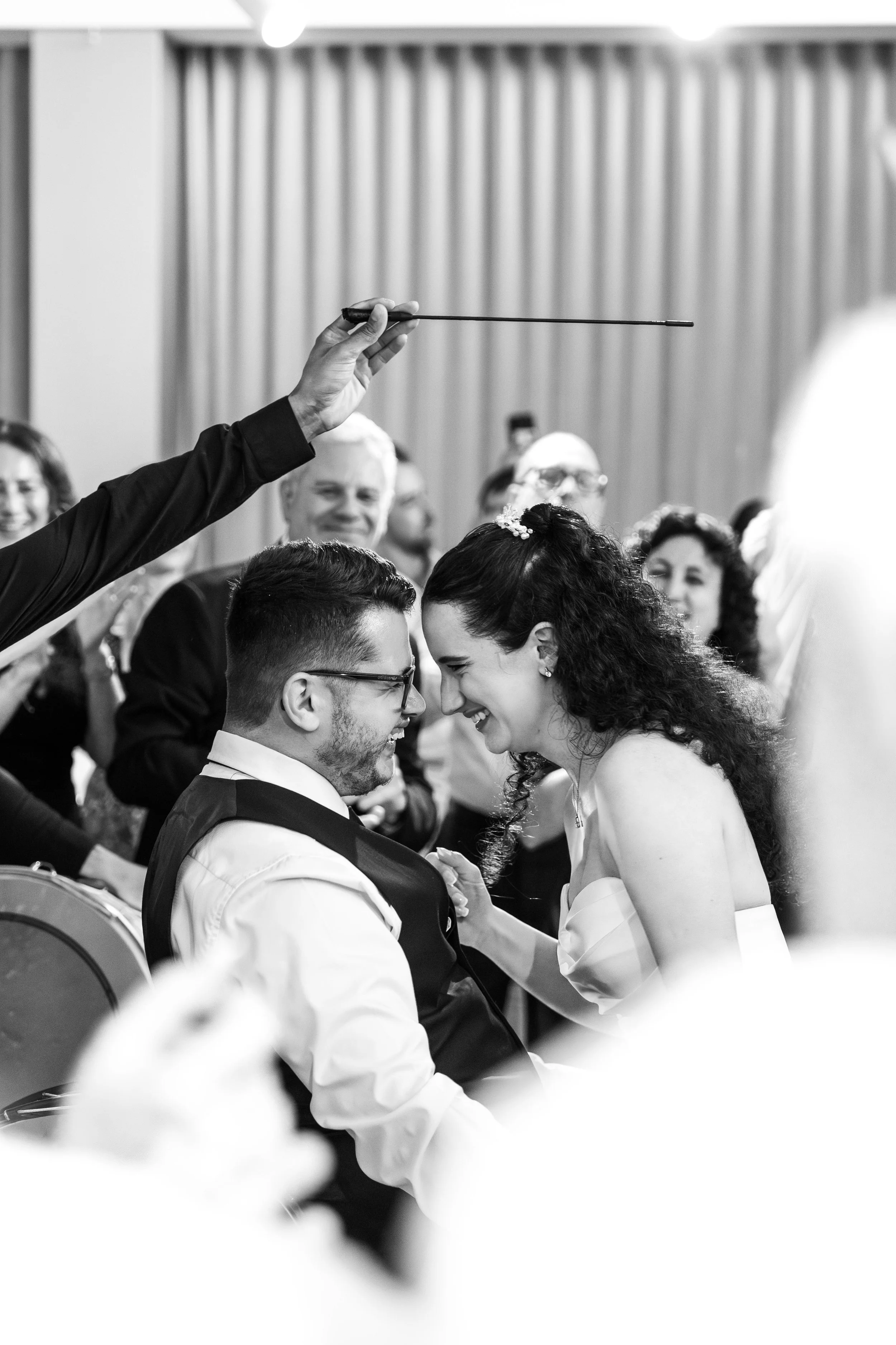Black and white photo of a wedding ceremony where a bride and groom are leaning their foreheads together and smiling, with guests and an officiant holding a thin stick above them.