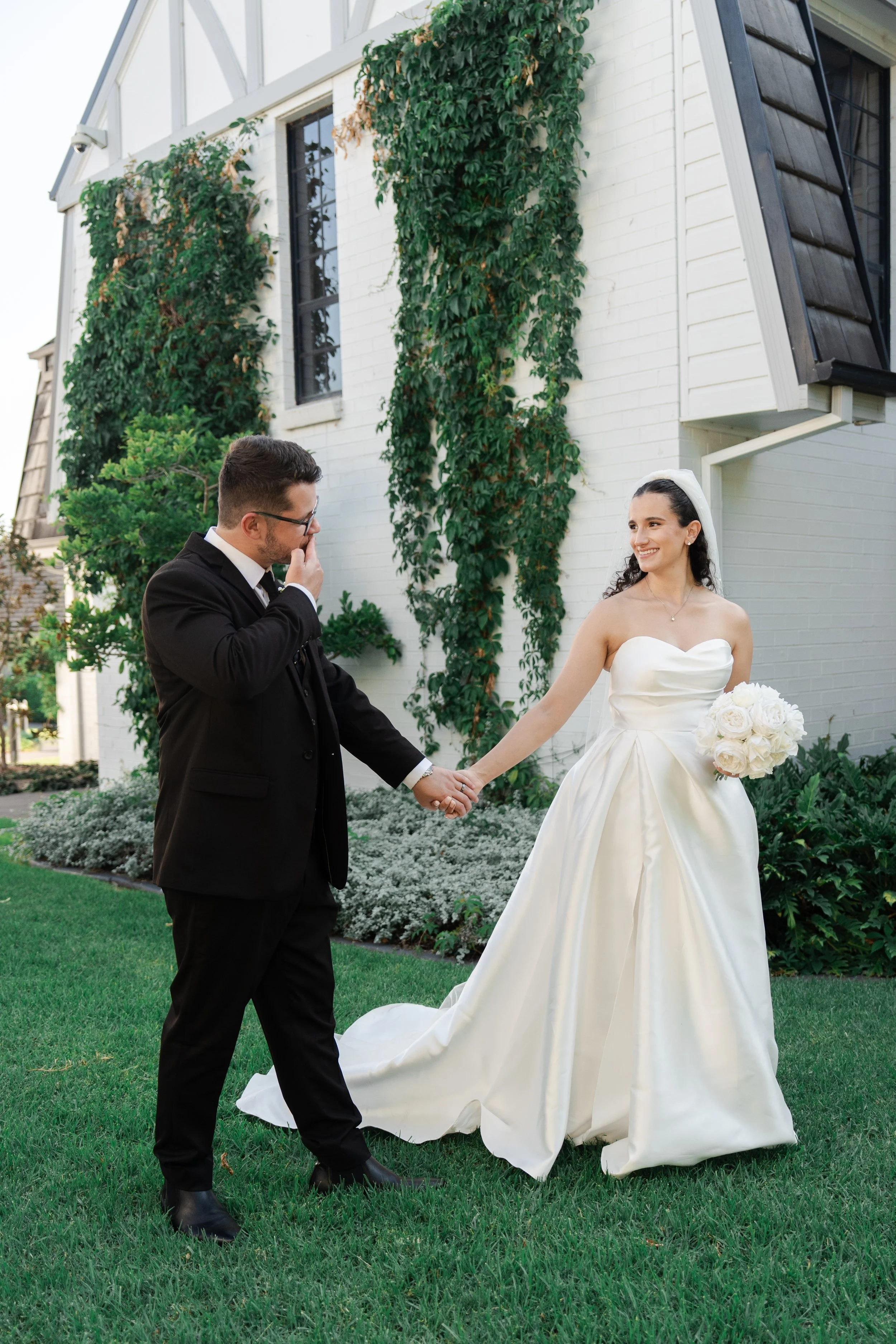 A bride in a white wedding gown holding a bouquet of white roses is holding hands with a groom in a black suit. They are outside in front of a white brick building with green vines and plants, during their wedding.