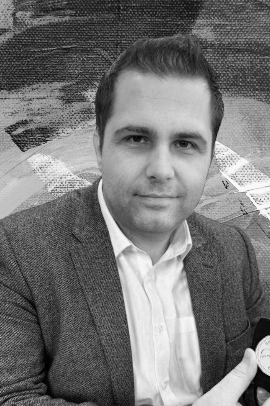 A black and white photo of a man in a suit with a white shirt, smiling subtly at the camera, with a background featuring artwork or textured patterns.