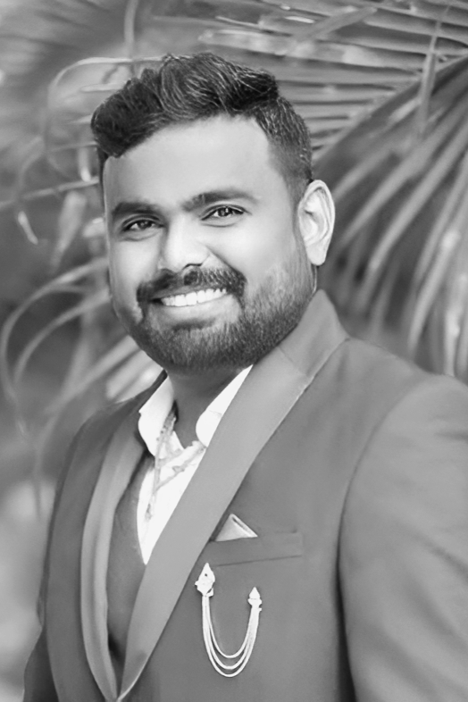 A man with dark hair and a beard, smiling, wearing a suit with a patterned shirt and a pin on his lapel, standing outdoors with leafy plants in the background.