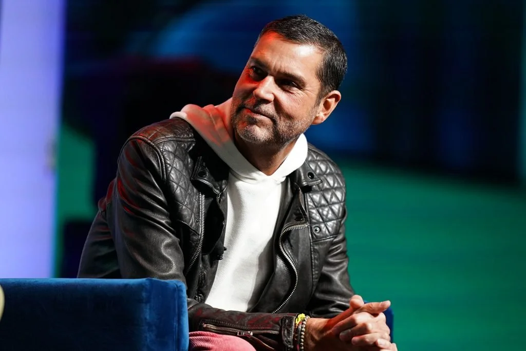 A man with short dark hair and a beard, wearing a black leather jacket over a white hoodie, sitting on a blue couch in a studio or stage setting with colorful lighting.