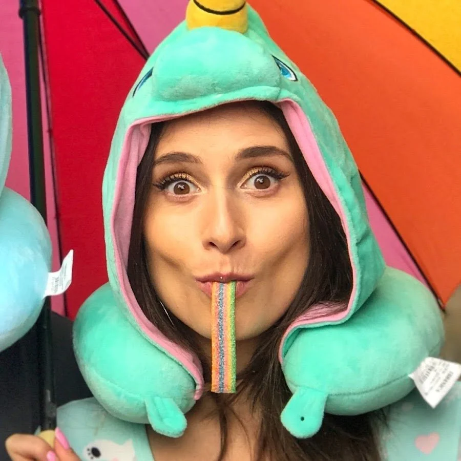 A woman is wearing a unicorn-themed hooded pillow that hugs her face and has a rainbow-colored candy in her mouth. The background is colorful and playful.