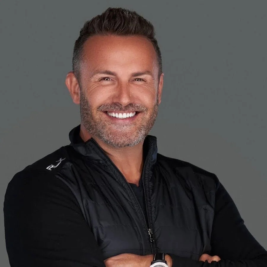 A smiling man with short gray hair, a beard, wearing a black Nike jacket, standing against a plain gray background.