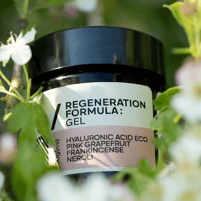 A black jar labeled 'elsewhere / Regeneration Formula: Gel' surrounded by green leaves and white flowers.