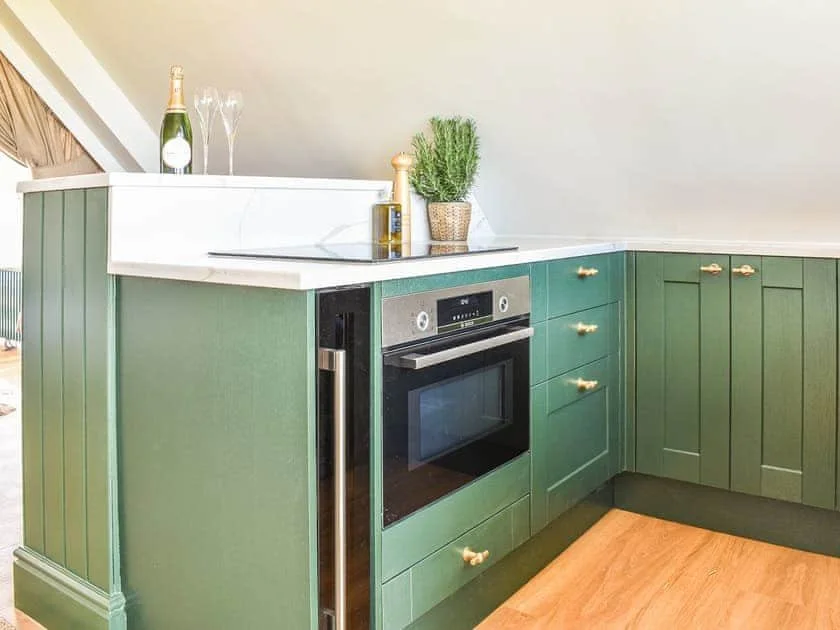 Modern kitchen with green cabinets, stainless steel oven, wood flooring, and marble countertop featuring champagne bottle, glasses, and potted plant.