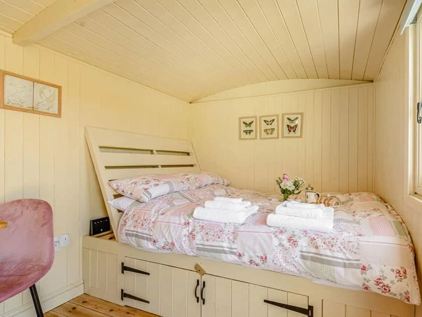 Cozy bedroom with a single bed featuring floral bedding, folded towels, a vase with flowers, and a cup on a tray. The room has wooden paneling and framed butterfly artwork on the walls.