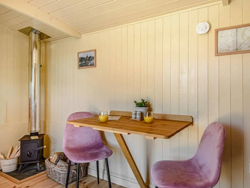 Cozy dining area with a small wooden table, two pink chairs, a wood stove, kindling basket, and a metal chimney pipe.