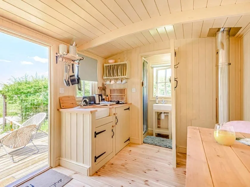 Cozy wooden kitchen interior with outdoor view, featuring a small stove, sink with rack, and adjacent bathroom. Natural light filters in through a window, and a dining table is visible with a glass of juice. Outdoors, there are chairs on a deck besid