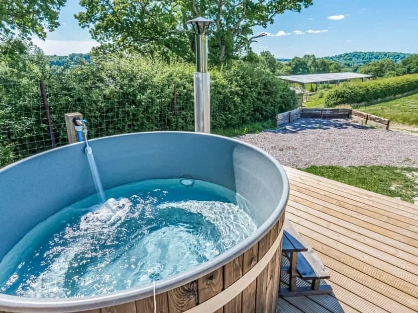 Outdoor wooden hot tub on a deck overlooking a scenic landscape with green fields and trees under a clear blue sky.