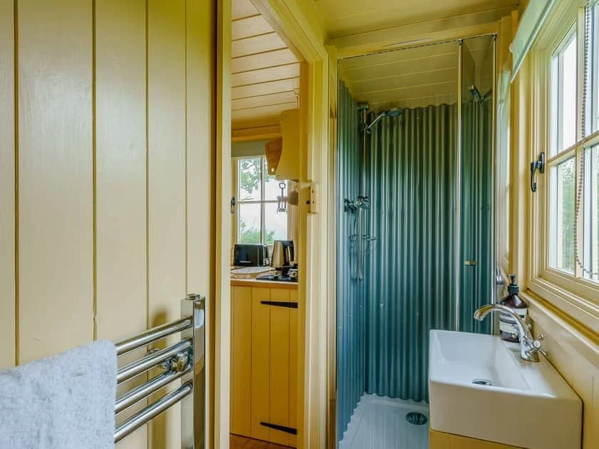 Compact bathroom with corrugated metal shower, small sink, towel rail, and view into adjoining kitchen with yellow cabinets.