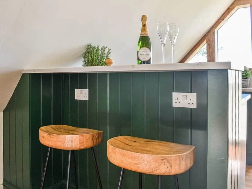 Bar area with green paneling, two wooden bar stools, a countertop holding a wine bottle, two glasses, and a small plant, under sloped ceiling with windows.