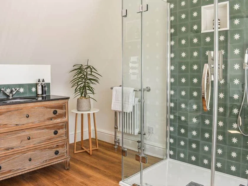 Modern bathroom with a glass shower enclosure, star-patterned green tiles, wooden vanity with a black countertop, and potted plant on a side table.
