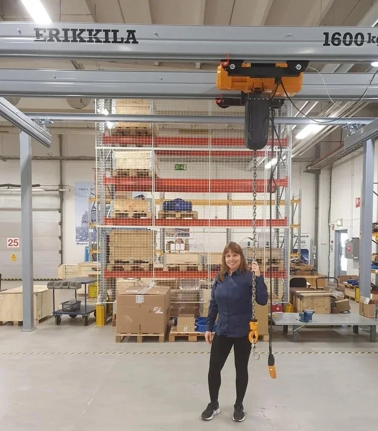 Nainen seisoo teollisuustilassa ja pitää kaapelinostinta, taustalla varasto ja hyllyjä