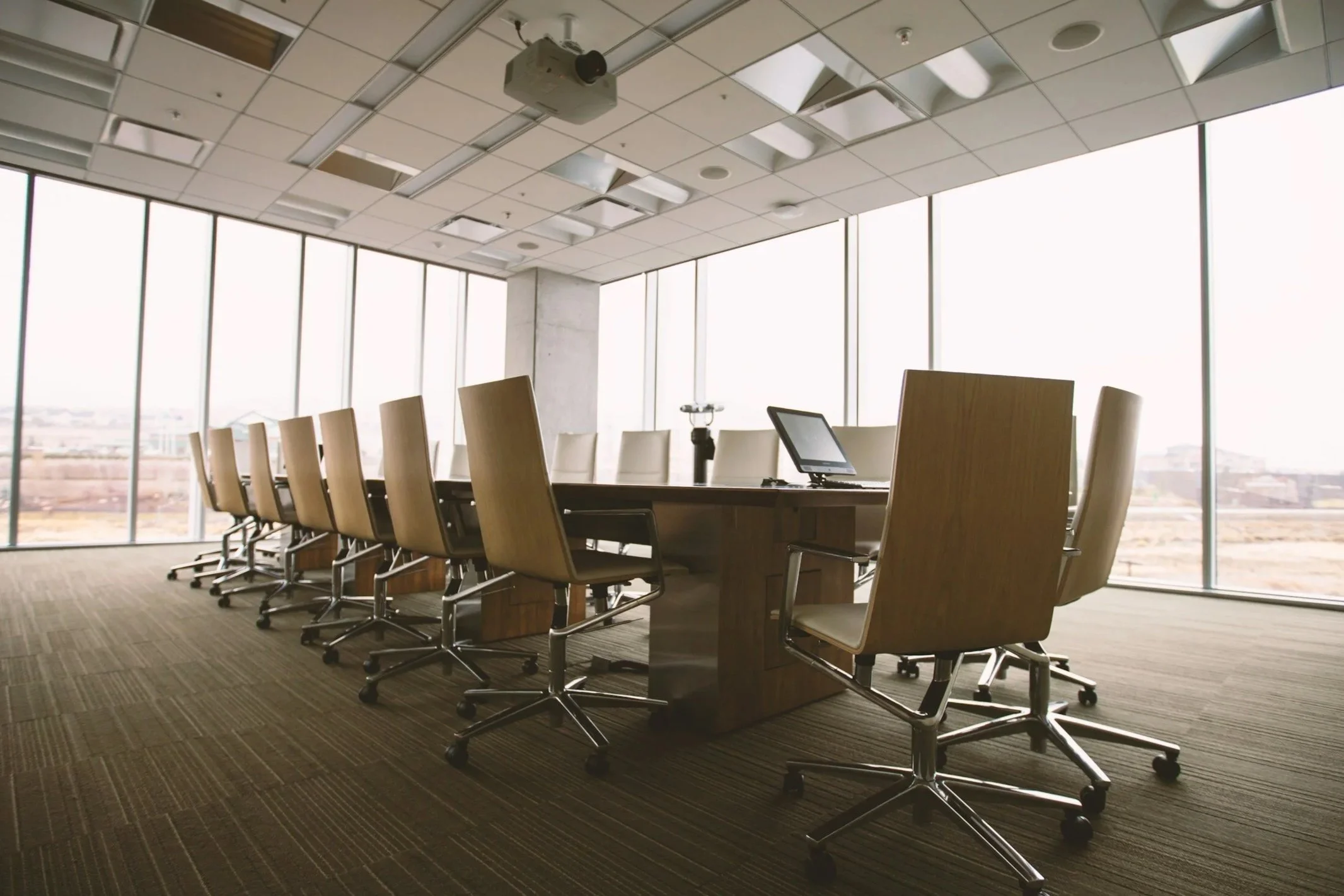 An empty conference room with a long wooden table surrounded by beige office chairs, a computer monitor on the table, large windows with city views, and a ceiling-mounted projector.