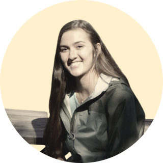 A young woman with long brown hair smiling, wearing a gray jacket, sitting with a yellow background.