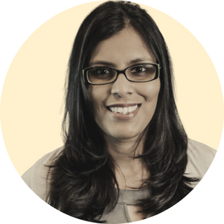 Sush Koka: A woman with long dark hair, glasses, and a light-colored top, smiling against a light background.