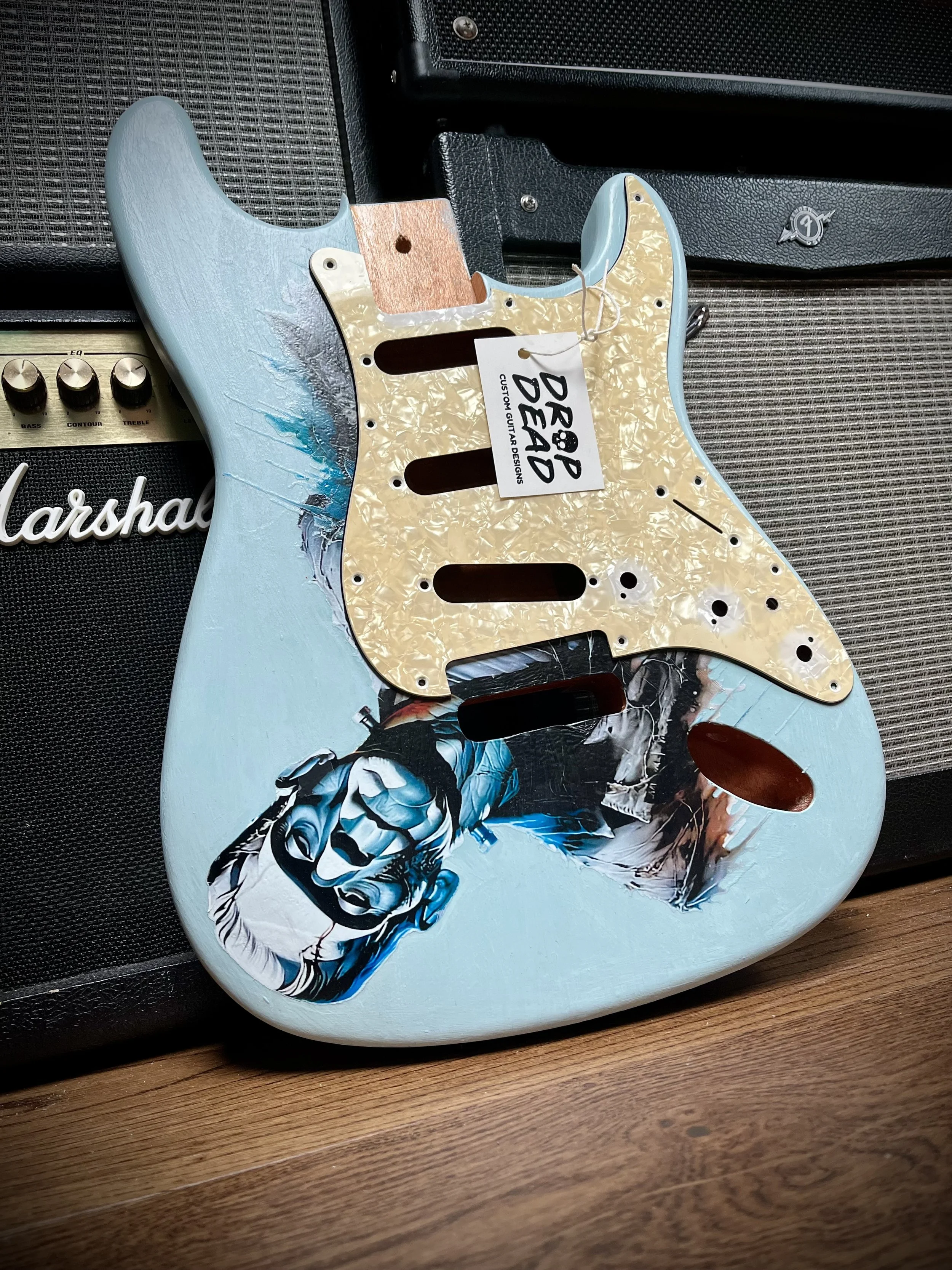 Electric guitar body with a detailed skeleton and floral artwork on a guitar pickguard, placed on a wooden surface with amplifier in the background.