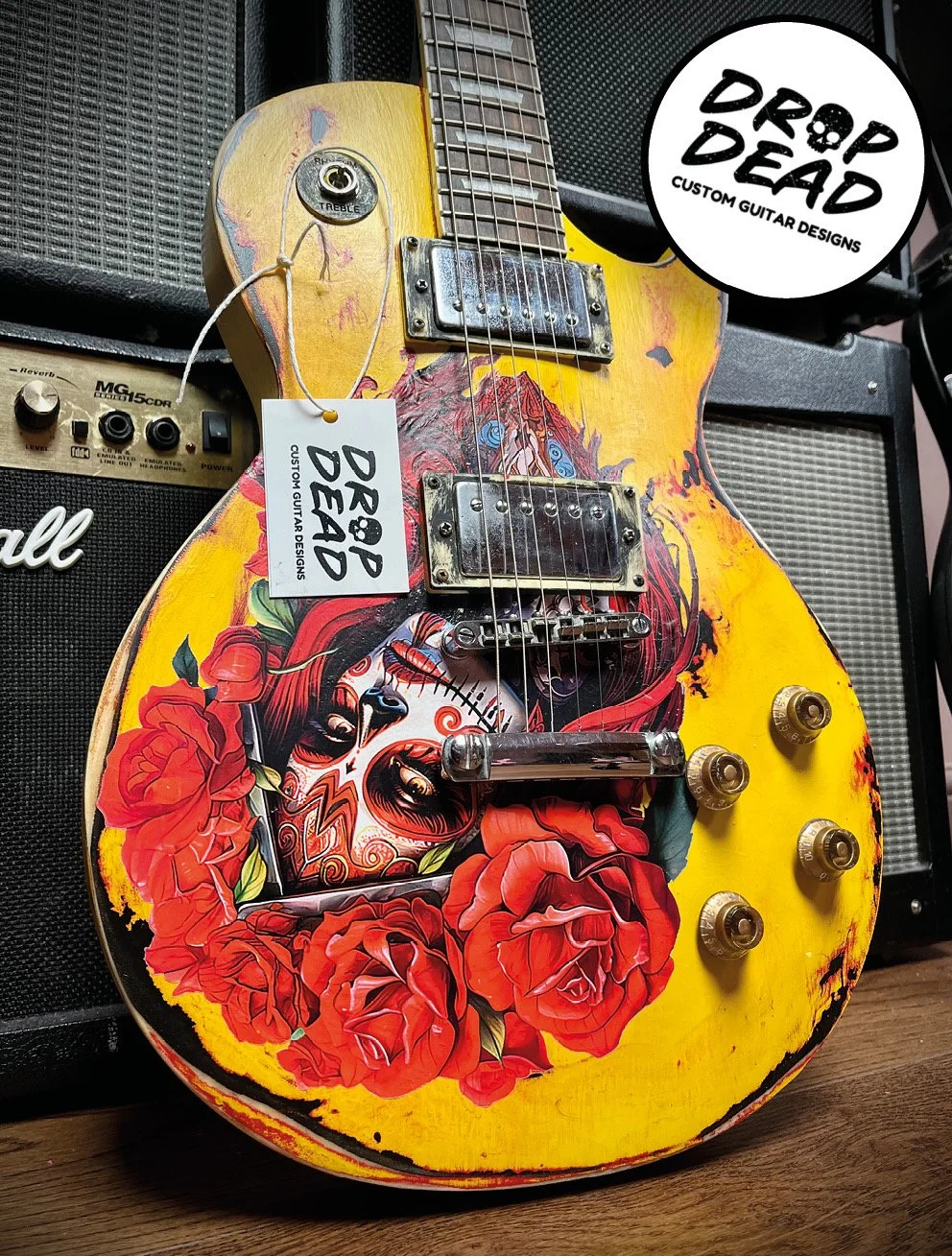 A yellow electric guitar with a colorful Day of the Dead skull and red roses painted on the body. The guitar has silver hardware and control knobs, with a white tag hanging from the neck that reads "Drop Dead Custom Guitar Designs."