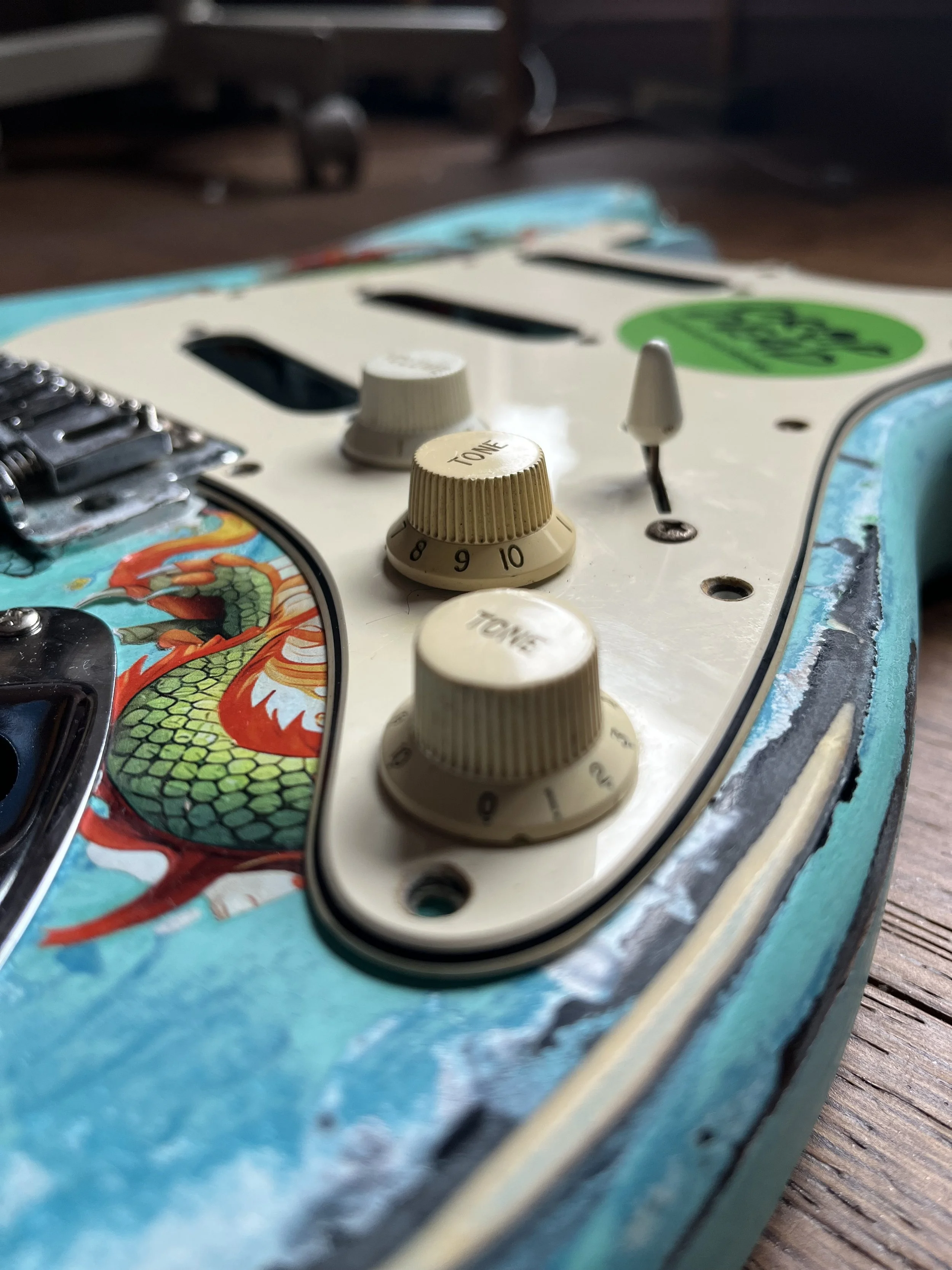 Close-up of a turquoise electric guitar with tone dials, a switch, and decorative dragon artwork.