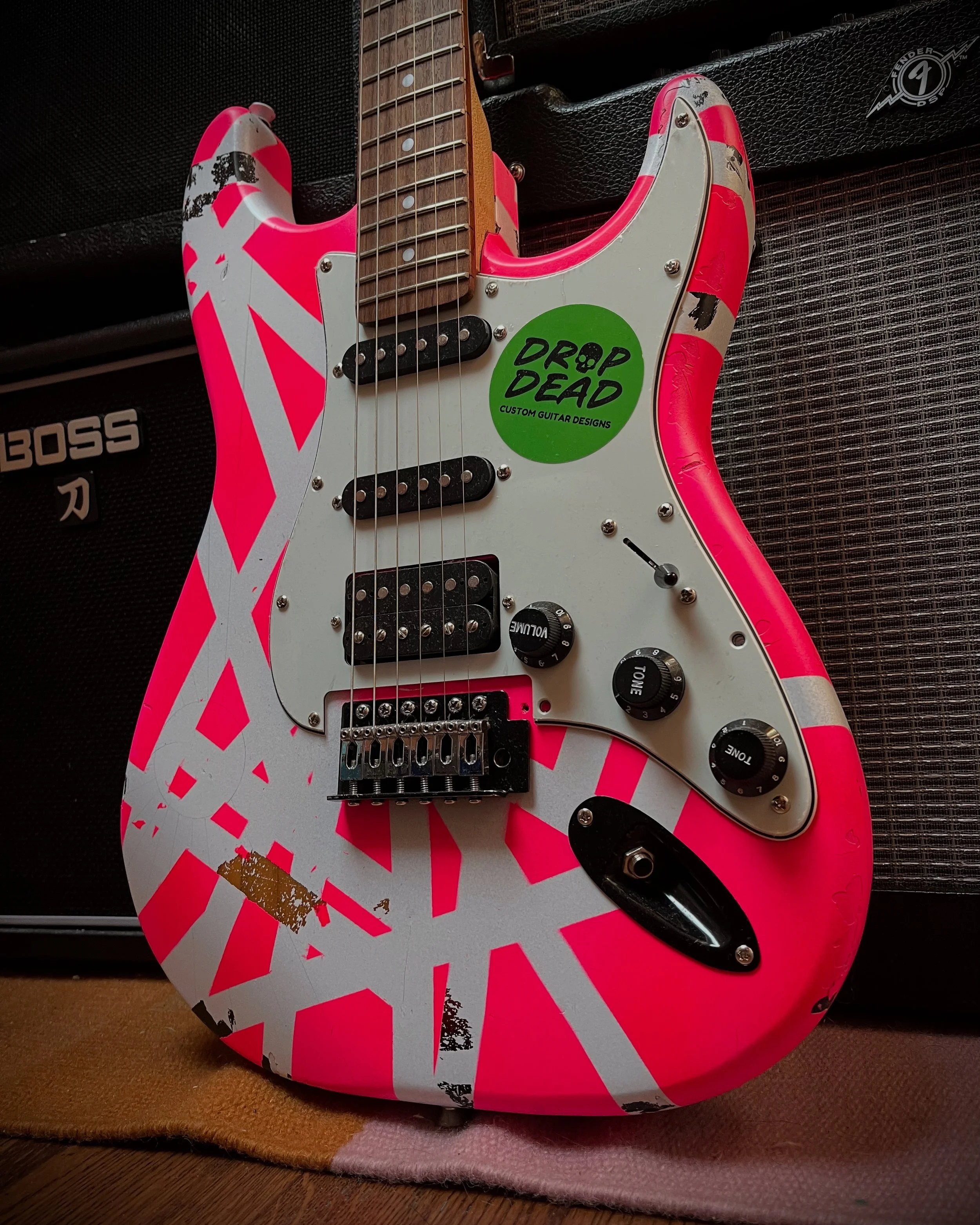 Pink and white electric guitar with a sticker that says "Drop Dead Custom Guitar Designs" leaning against an amplifier.