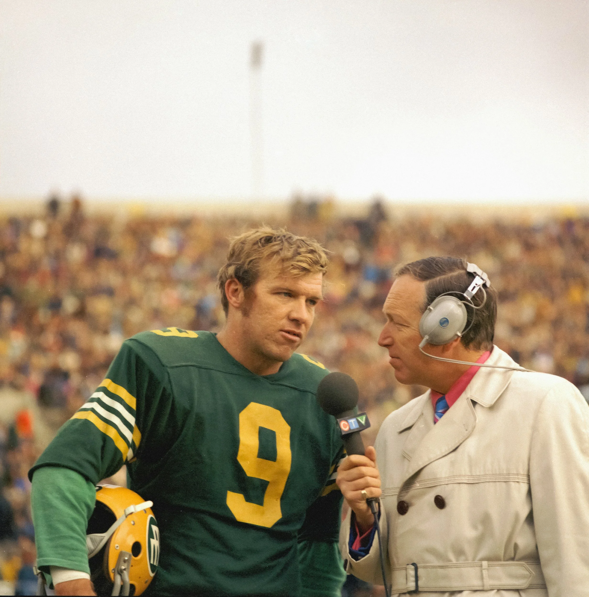 A football player in a green and yellow uniform with the number 9 on it is being interviewed by a sports announcer with a headset and a microphone at a football game, with a large crowd in the background.