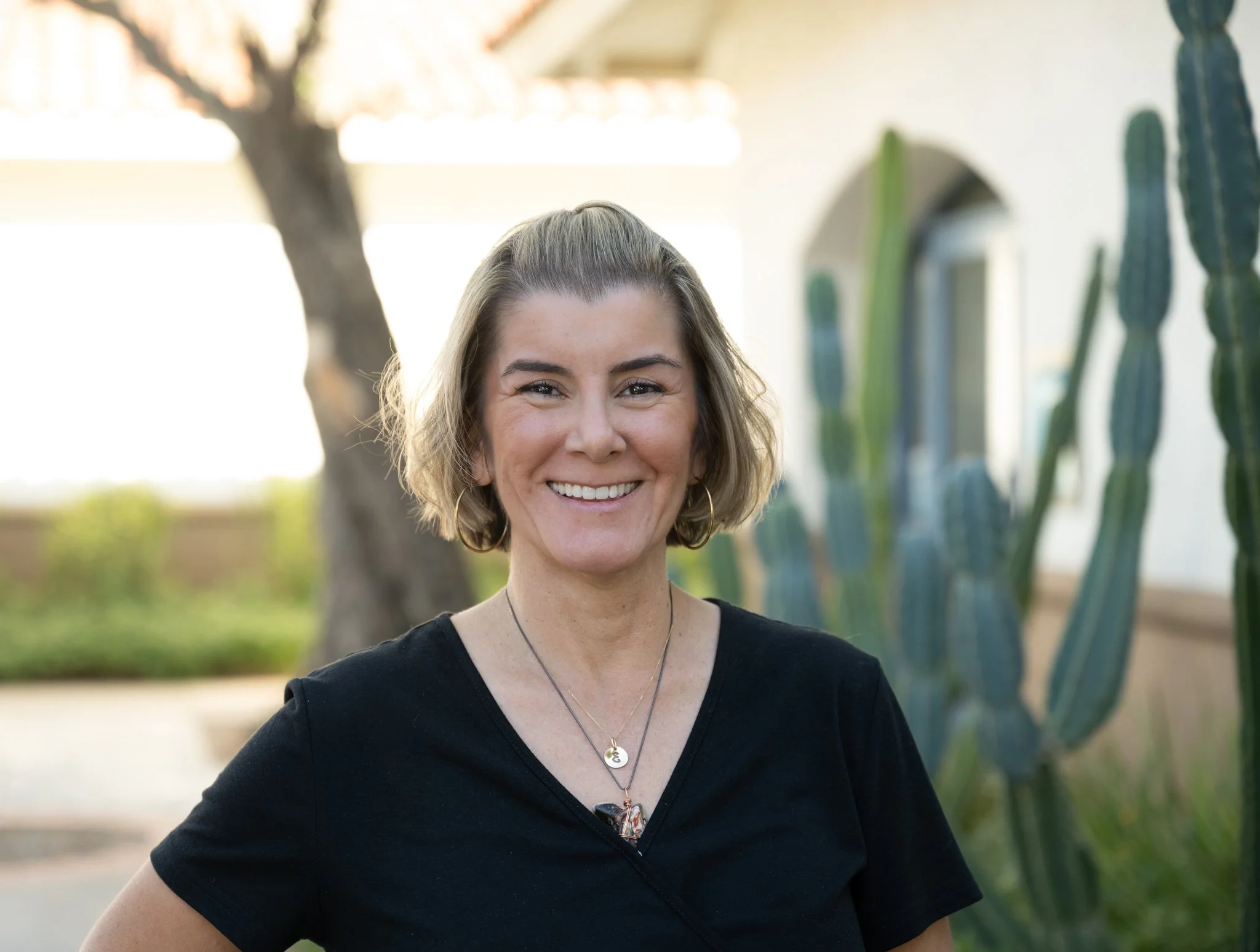 Smiling woman with short light brown hair wearing a black top and necklaces, standing outdoors with cacti and trees in the background.