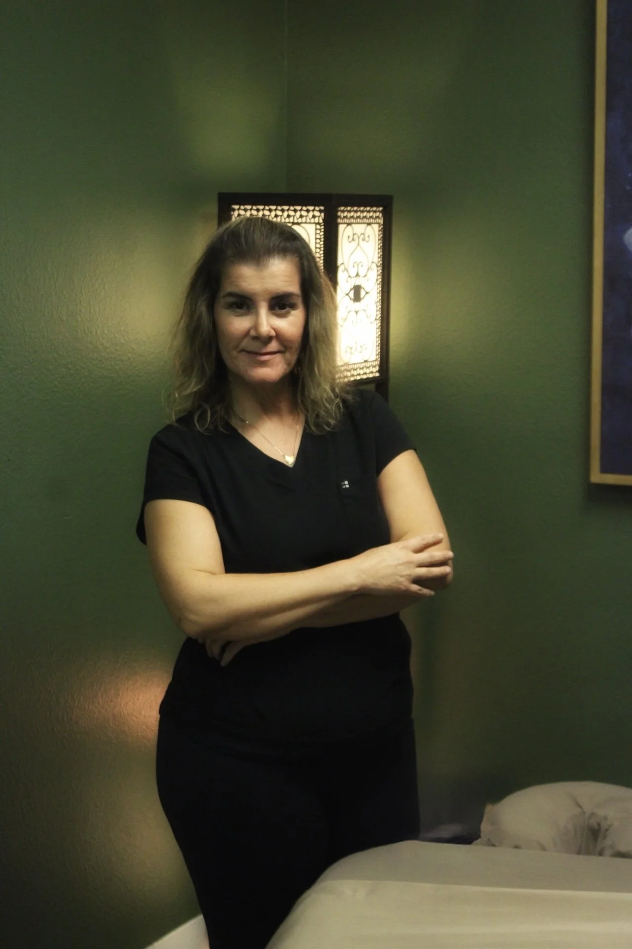 A woman with wavy brown hair standing in a room with dark green walls. She is wearing a black t-shirt and black pants, with her arms crossed. Behind her, there is a decorative light fixture with a face design, illuminating the wall.