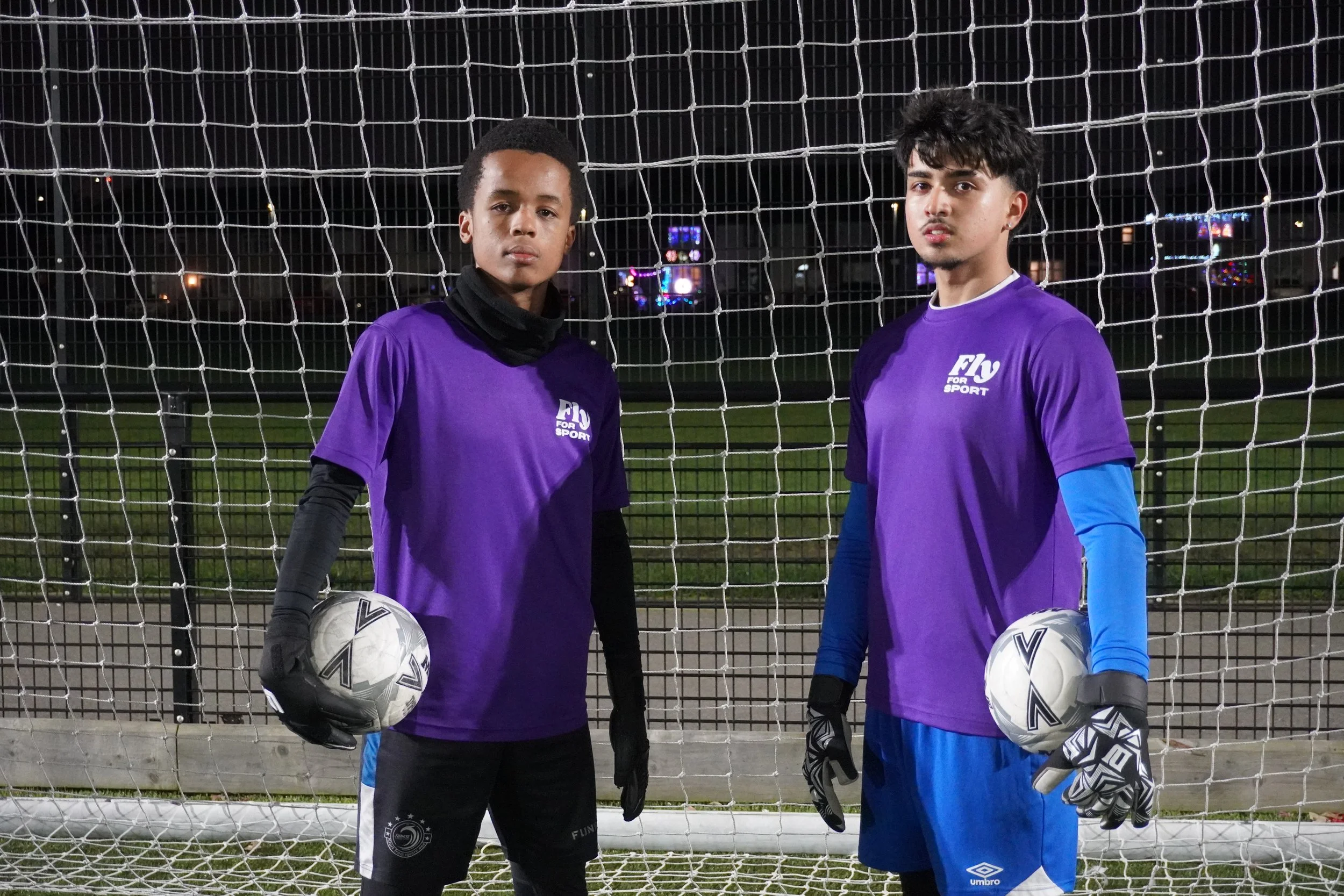 Two young male soccer players standing in front of a goal on an outdoor soccer field at night. They are wearing purple jerseys, with one holding a soccer ball in each hand. The player on the left is wearing black gloves and a black neck warmer, while the player on the right is wearing black gloves and a long-sleeve blue undershirt.