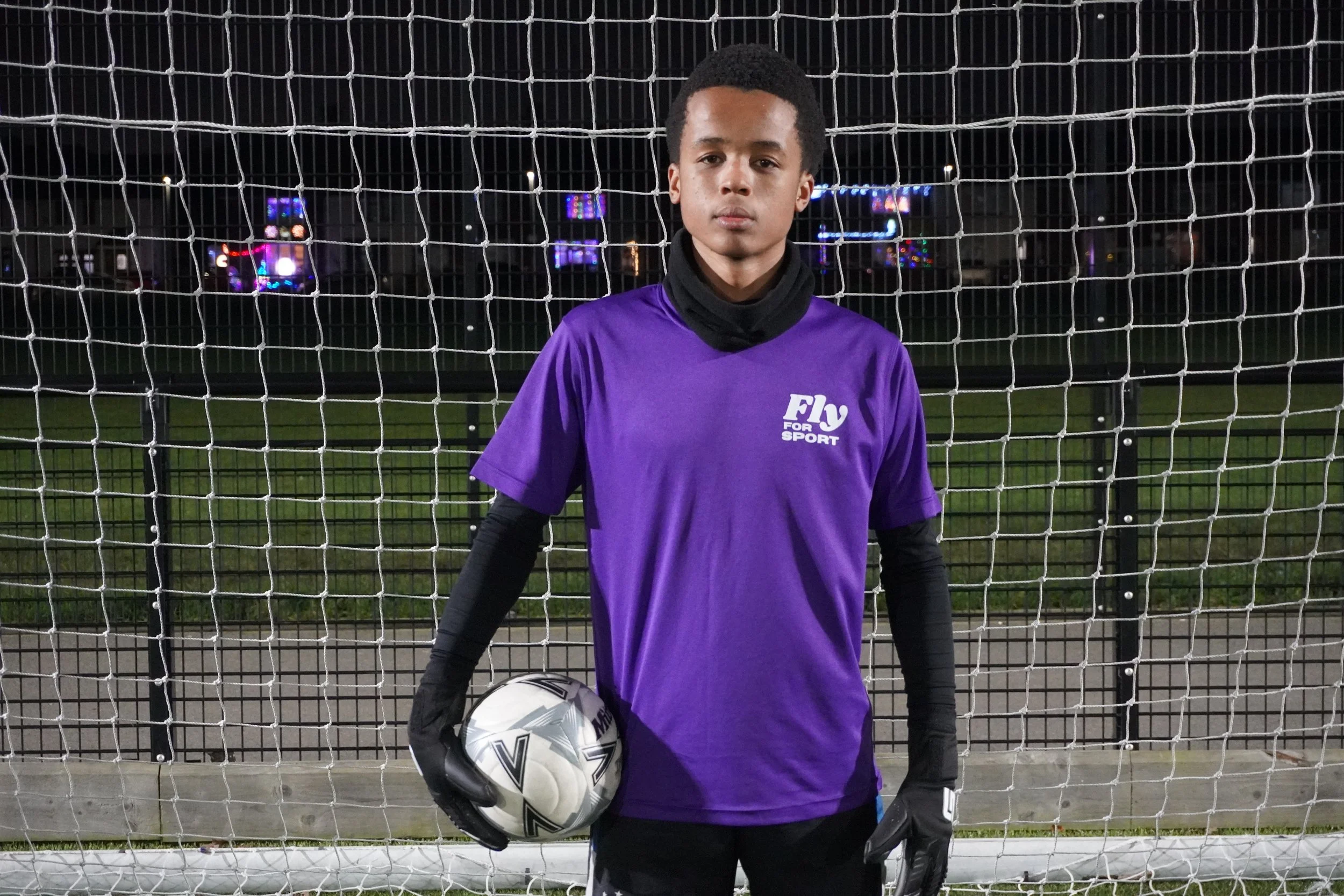 A young male soccer goalkeeper standing in front of a goal net at night, holding a soccer ball, wearing a purple sports jersey and black gloves.