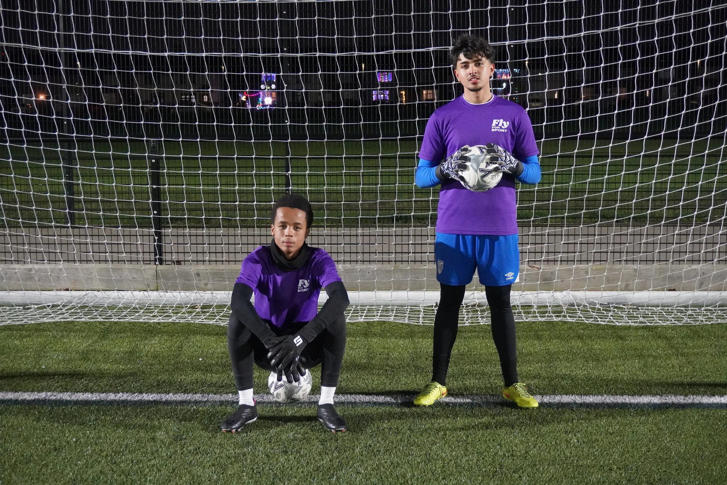 Two young male soccer players in purple jerseys stand in front of a soccer goal on a field at night, one squatting with a soccer ball at his feet, the other standing holding another soccer ball. Both wear black gloves and black or yellow shoes.