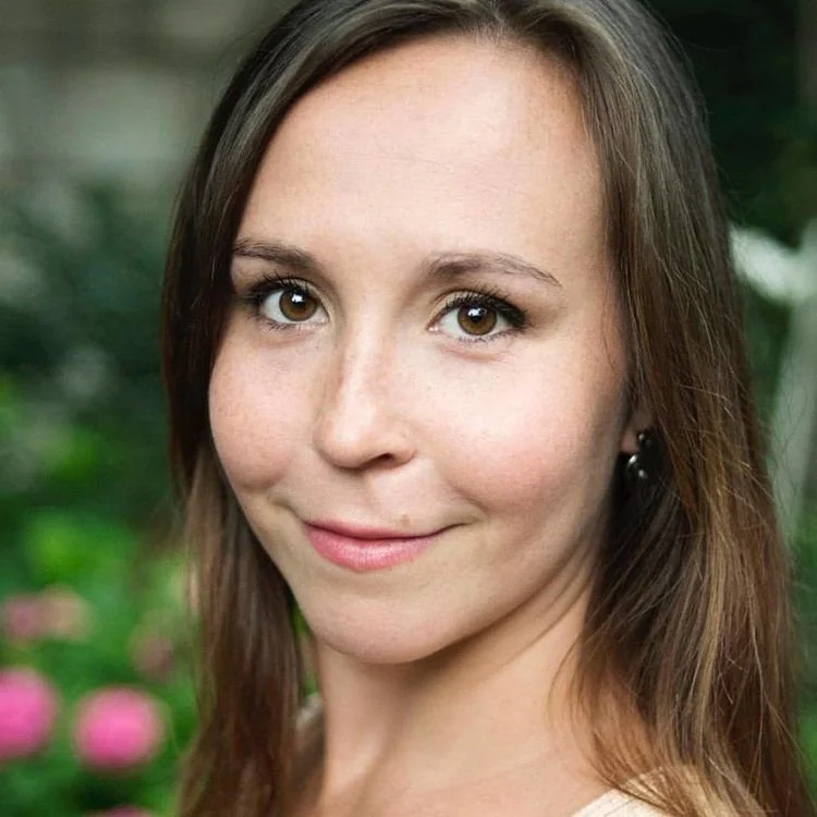 Close-up portrait of a young woman with brown hair and brown eyes, smiling softly, with a blurred green garden background.