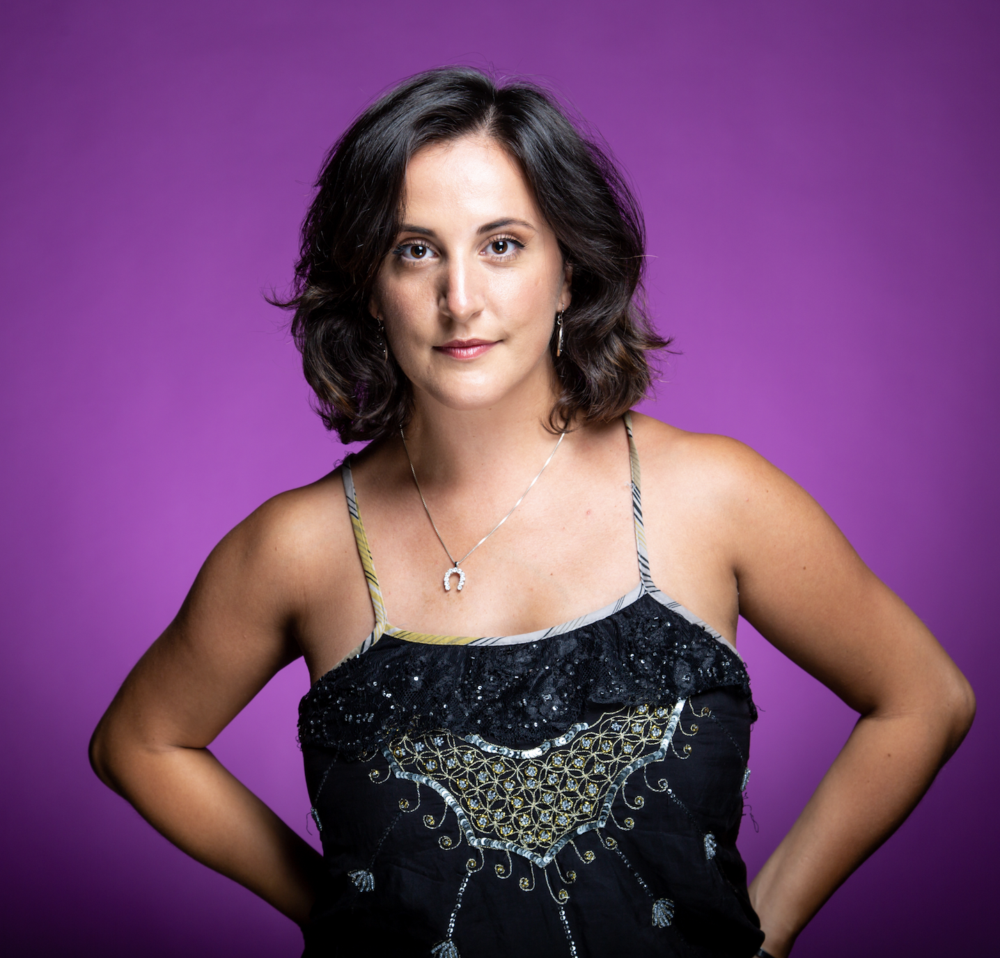 A woman with shoulder-length dark hair wearing a black sequined top and a silver necklace with a horseshoe pendant, standing against a purple background.