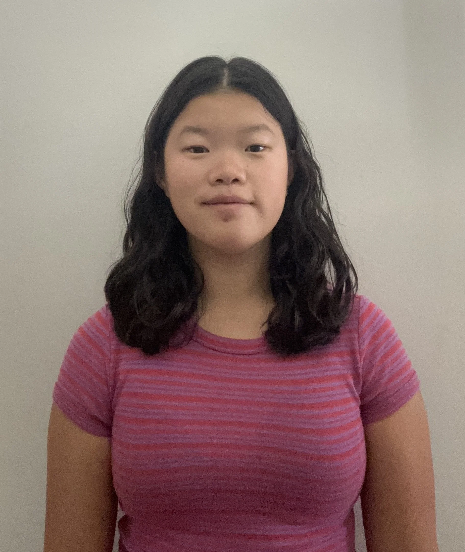 A young girl with shoulder-length curly black hair, wearing a pink and purple striped t-shirt, standing against a plain beige wall.