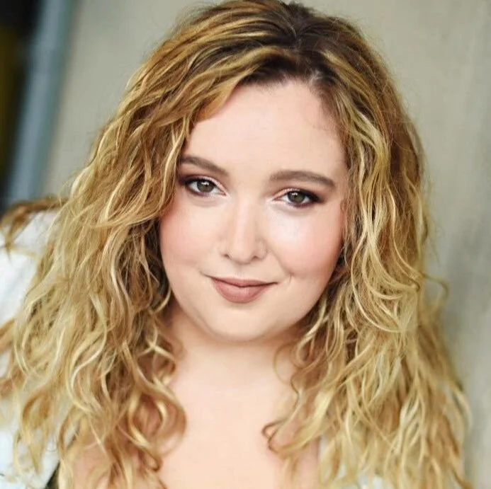 A woman with long, curly blonde hair and light makeup, smiling softly at the camera.