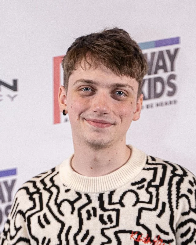 Portrait of a young man with short brown hair, wearing a red and white striped shirt, against a plain blue background.