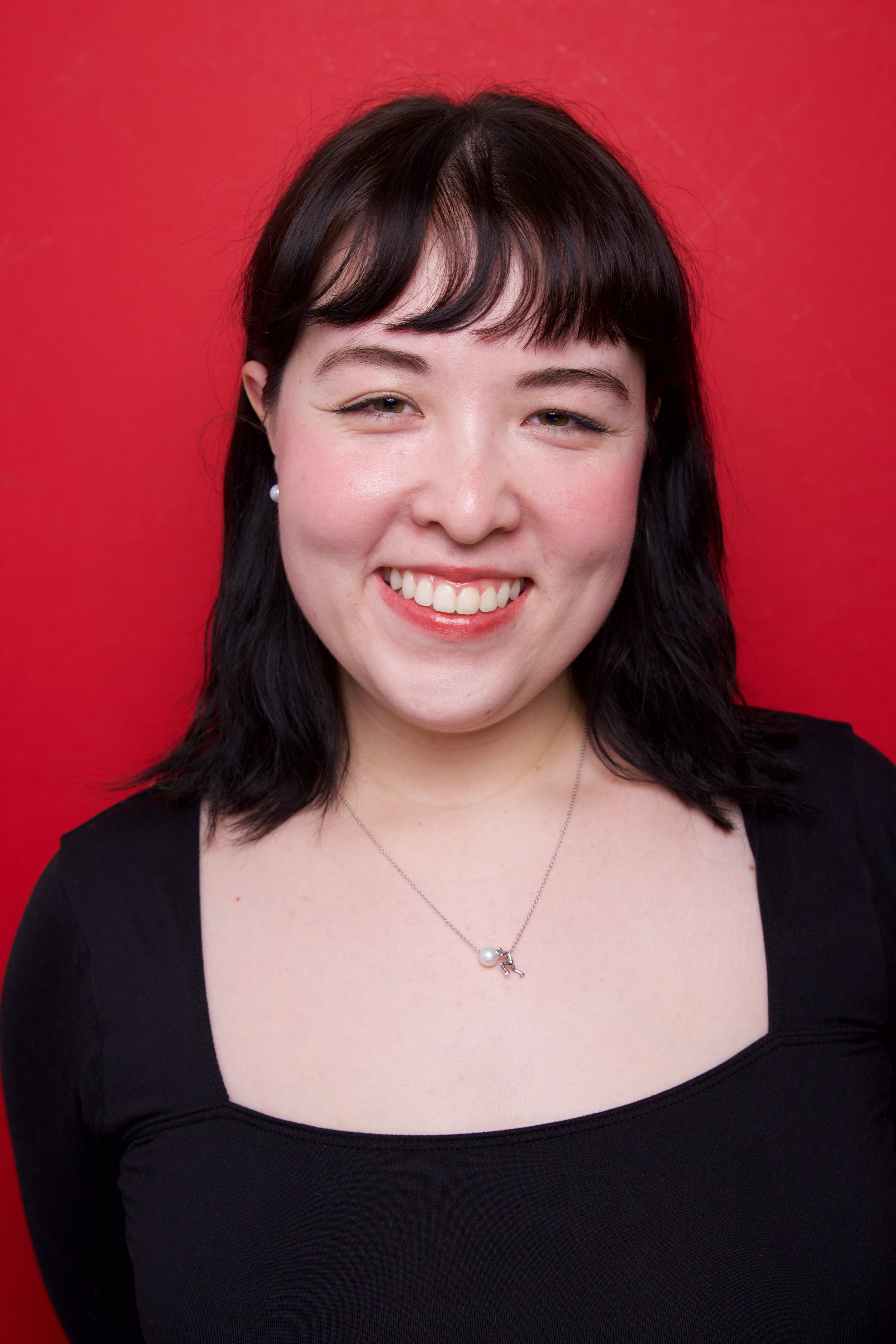A woman with short dark hair, wearing black shirt and a necklace, smiling against a red background.