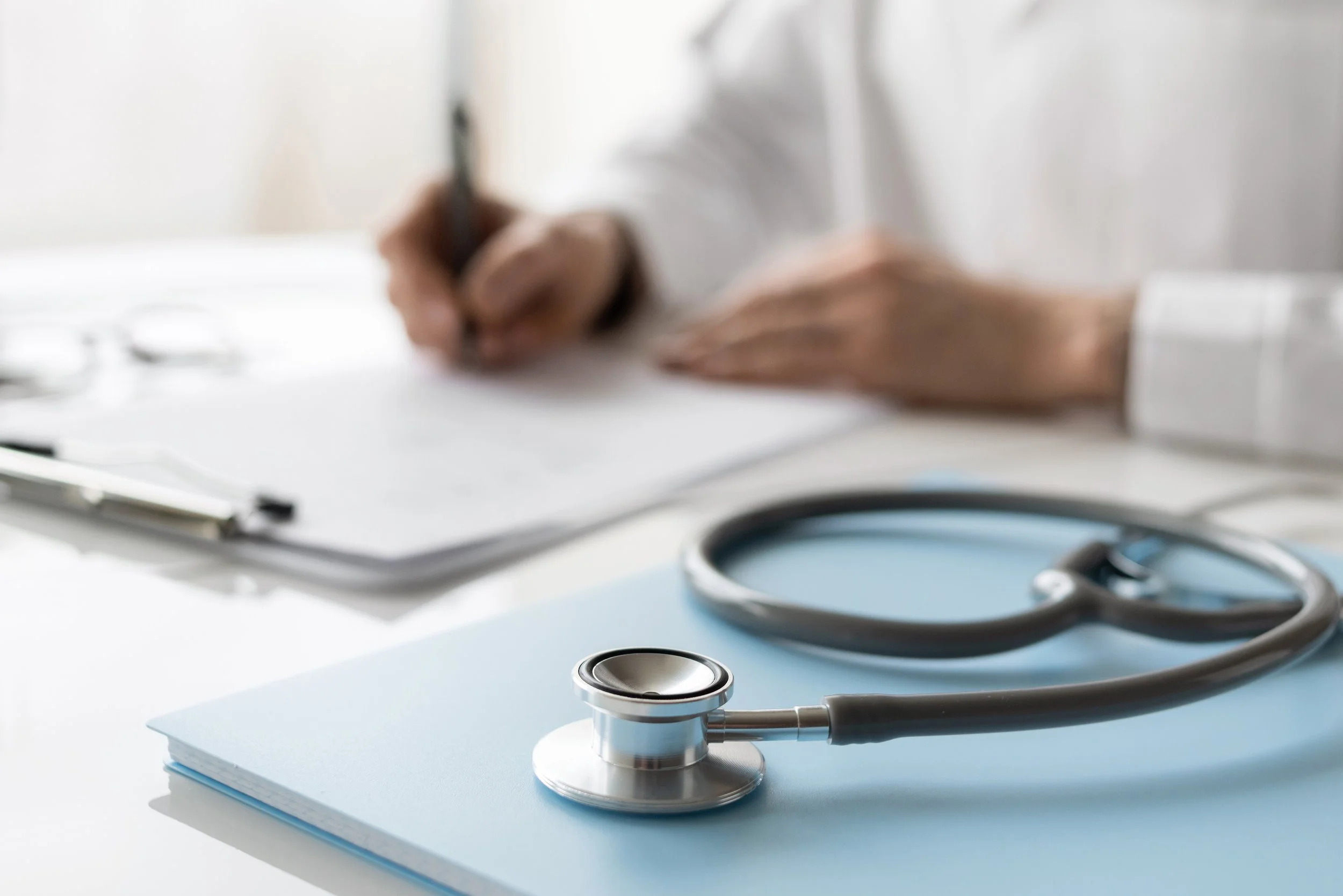 A physician's hand writing on a piece of paper on a clipboard, next to it a notebook with a stethoscope on it